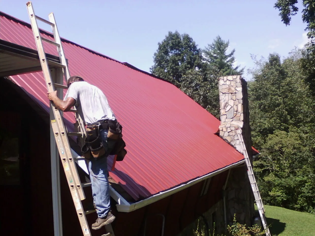 Roofer on a ladder beside a red metal roof and stone chimney — Soffit & Fascia Repair by Roofing Brothers in Crivitz